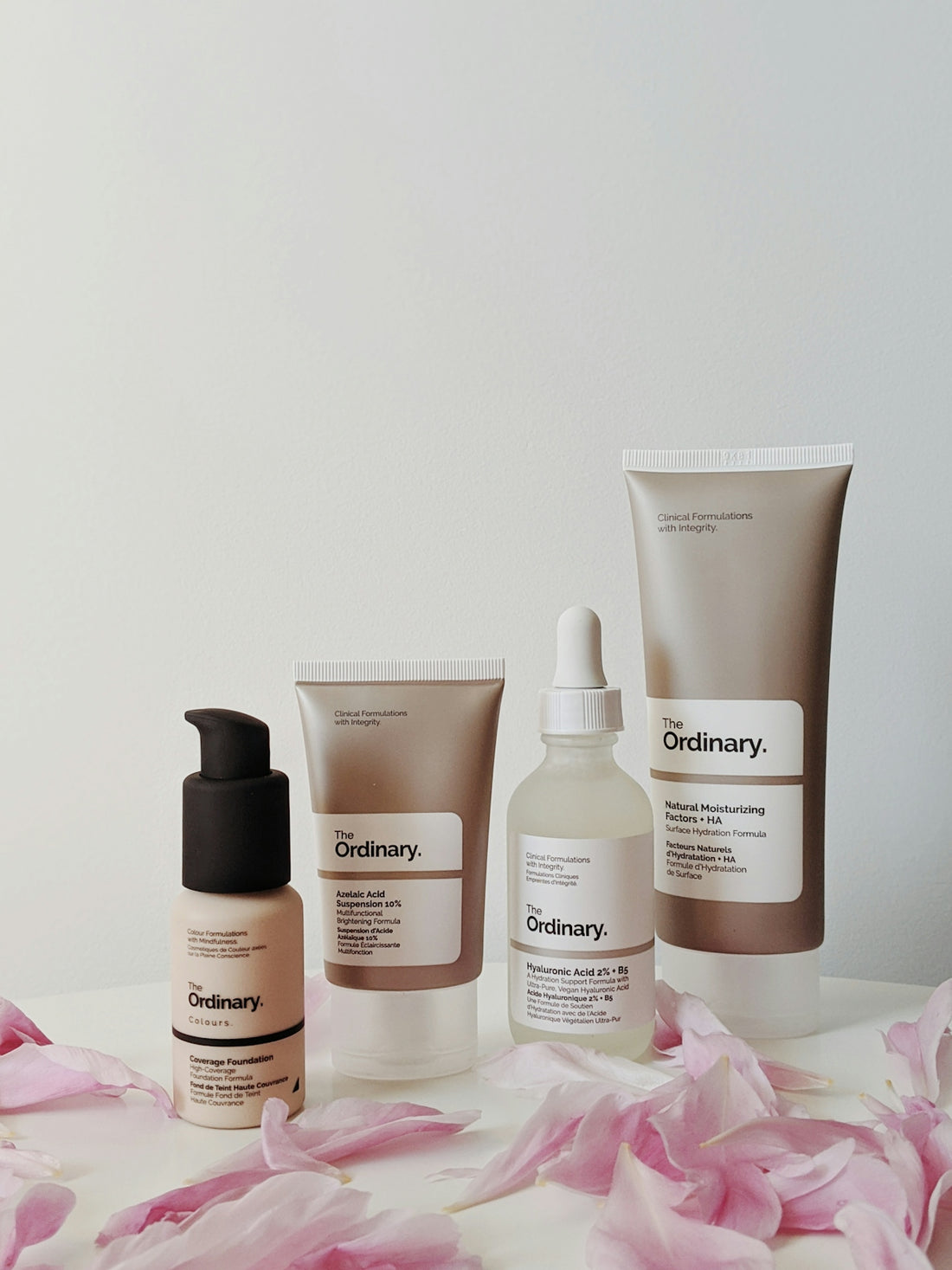 Four skincare products from The Ordinary arranged with pink flower petals on a table