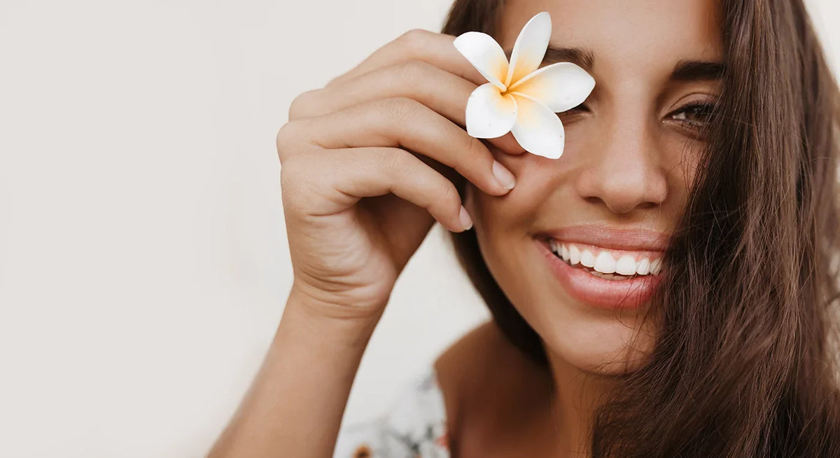 Smiling woman holding a white flower over one eye against a light background.